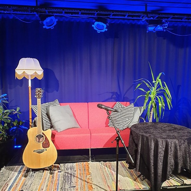 Bild på en scen med soffa, golvlampa, blommor, gitarr och en mick