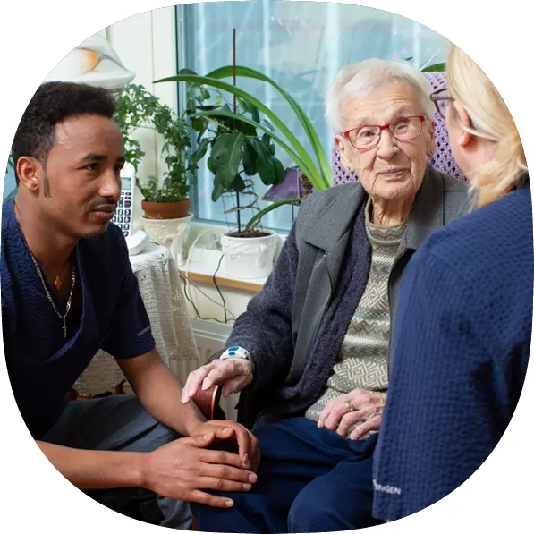 En äldre person sitter tillsammans med två personer som arbetar med omsorg i hemmiljö.