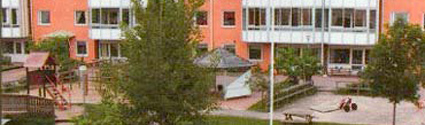 Förskolan Dag och Natt ligger i ett hyreshus med rödbrun fasad och vita balkonger. Framför förskolan finns en förskolegård med lekredskap och flera träd