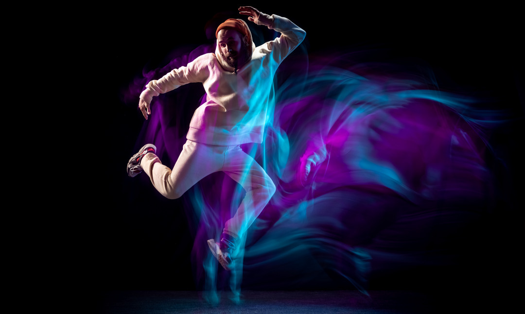 Jumping. Energy young sportive man dancing hip-hop or breakdance in white outfit on dark background in mixed blue neon light. Concept of sport, art, action, moves, youth culture. Banner for ad