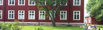 Rött hus där Trollgårdens förskola ligger. Framför huset står ett stort träd och en grön gräsmatta.