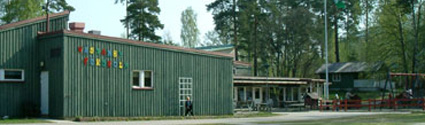 Grönt trähus med texten Västanby förskola på fasaden. Bredvid huset finns flera träd.