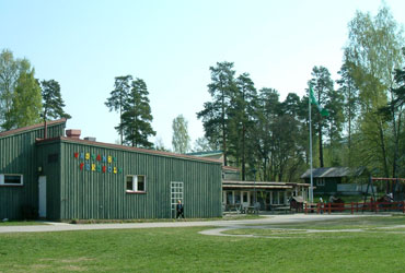 Grönt trähus med texten Västanby förskola på fasaden. Bredvid huset finns flera träd.
