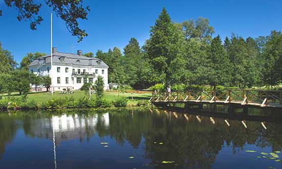 Nya herrgården i Högbo som speglas i vattnet. En gångbro leder ut i vattnet och i bakgrunden finns skog mot en klarblå himmel.