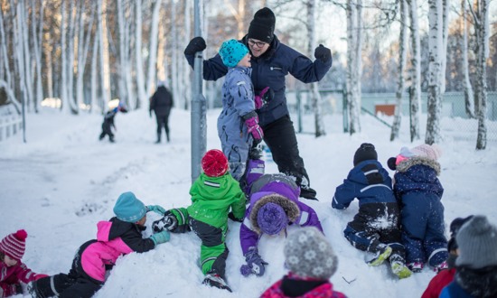 Flera förskolebarn kryper omkring på en snöhög, på toppen av snöhögens står ett barn och en pedagog och ropar tillsammans.