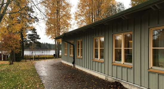 Alsjöskolan ligger i en enplansfastighet med grön träfasad. Bakom skolan syns träd och en sjö.