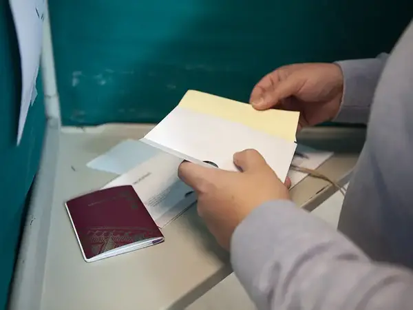 En hand håller ett valkuvert. Ett rött pass ligger bredvid på bordet.