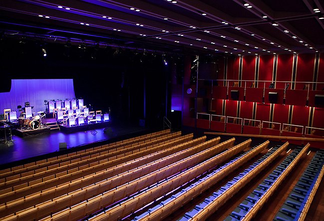 Teatersalong med tomma bänkrader och en rödfärgad bortre vägg. På scenen finns ett trumset och platser för musiker som lyses upp av lilafärgade strålkastare.