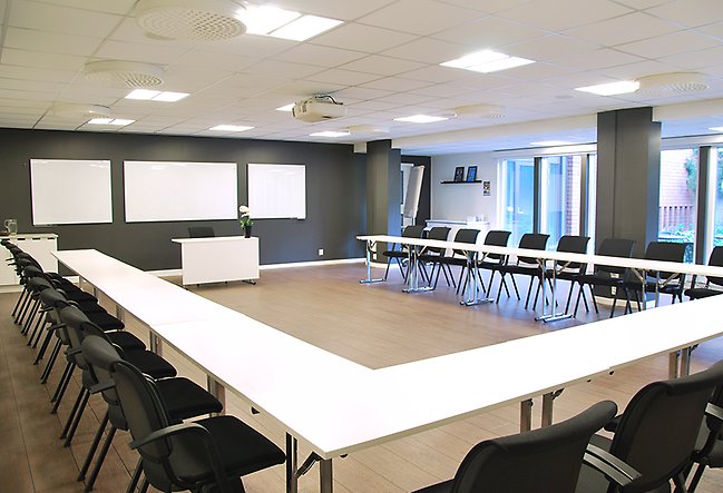 Stor konferenslokal med bord format som ett U. Runt bordet finns svarta stolar. Längst fram i lokalen finns skrivtavlor på väggen och den andra väggen som syns består av fönster.